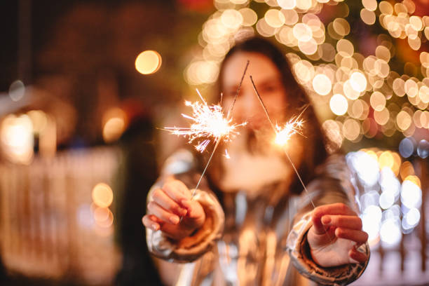 Young happy woman holding sparklers while standing against illuminated Christmas tree outdoors in city stock photo