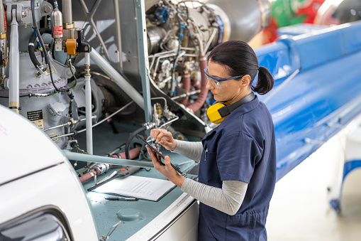 Female mid adult aviation mechanic standing near helicopter engine and looking at digital tablet, side view. Engineer in protective workwear fixing air vehicle motor in hangar, medium shot