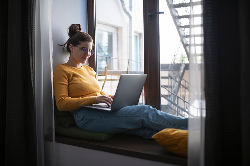 Young woman sitting at the window at home using laptop.