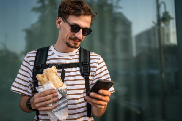 man caucasian tourist young adult eat sandwich and use mobile phone stock photo