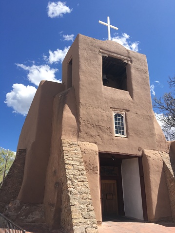 San Miguel Church in Santa Fe, New Mexico. Oldest Catholic Church in the United States, built in the 15th Century. Adobe style, rock, one window, bell tower, simple white cross atop church. Blue sky with some fluffy white clouds.