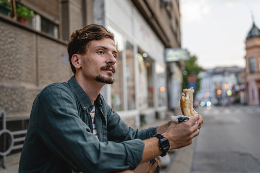 One man young adult modern caucasian male in the city in sunny day stand and eat sandwich fast food concept urban life copy space tourist eating in front of building wall real person