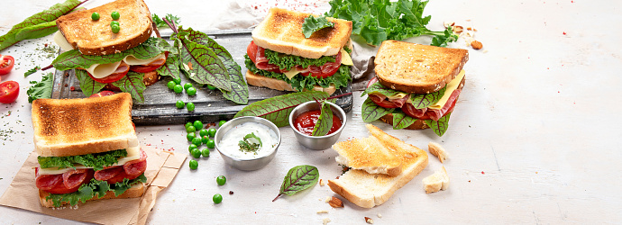 Sandwich with cheese, ham, tomato and fresh salad, made of whole wheat bread with a crispy crust on a wooden board with sauces on a white background, top view, panorama with copy space.