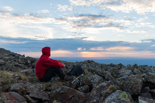 A man in a red jacket with a backpack and trekking poles enjoying the sunset at the top of the mountain. rear view. Hiking concept. Travel around the country.