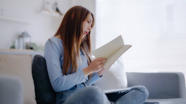 istock Young asian woman reading a book while seated on couch at home 1433015582
