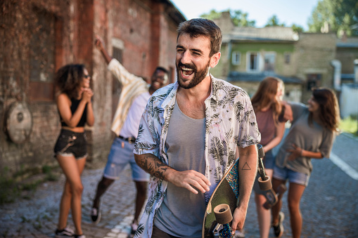 Cheerful hipster man holding his skateboard on the street with his friends in the background.