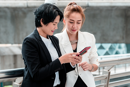 Two Asian woman talking business partners using digital tablet smartphone meeting outside office at street city. Teamwork partnership women talking discuss at outdoor office partnership team advice.