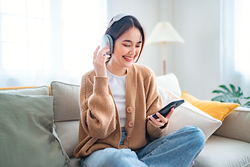 Happy young asian woman relaxing and listening music and watches movie on mobile phone with headphones while sitting on a couch in the morning at home