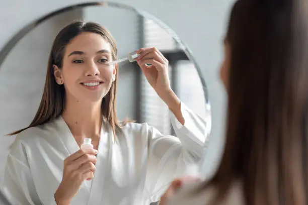 Skin Care Concept. Beautiful Millennial Woman Applying Face Serum Near Mirror, Happy Young Female Standing In Bathroom, Making Daily Skincare Routine At Home, Selective Focus On Reflection