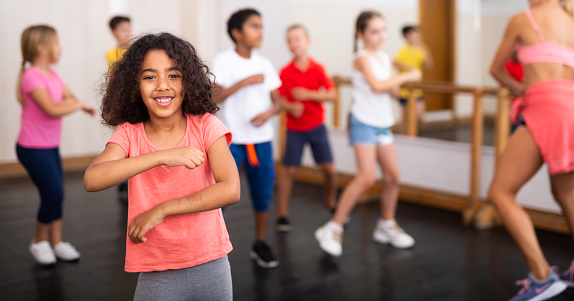 Cute smiling mulatto preteen girl exercising with group of children in choreography class