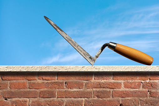 Close-up of a dirty concrete trowel with wooden handle above a brick and marble wall with blue sky on background, photography.
