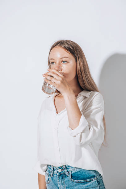 Girl drinks water from a glass cup on a white background. The concept of maintaining water balance stock photo