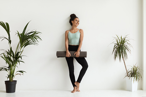 Portrait Of Sporty Young Female In Activewear With Fitness Mat In Hands Standing Near White Wall In Spacious Room, Smiling Fit Millennial Woman Ready For Yoga Or Pilates Class, Copy Space