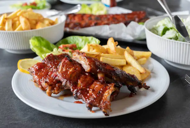 Delicious homemade oven baked spareribs with french fries and salad. Served ready to eat on a dinner table. Front view with blurred background