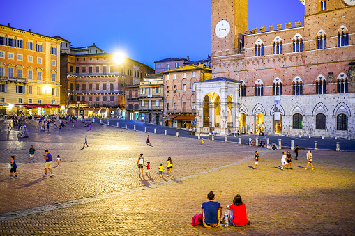 Siena, Tuscany, Italy, July 07 -- A couple enjoys a summer evening sitting on the pavement of Piazza del Campo, in the medieval heart of Siena, with the Palazzo Pubblico in background. Built starting in 1290 for the seat of the Council of Nine and the executive power of the Sienese city, the majestic Palazzo Pubblico is still the seat of the municipal government. Center of the city life since 1169, Piazza del Campo or simply Il Campo is one of the most beautiful and famous squares in the world for its particular shell shape. In this space every year the seventeen historical villages of Siena compete in the Palio, one of the oldest horse races in the world. Siena is one of the most beautiful Italian cities of art, in the heart of the Tuscan hills, visited for its immense artistic and historical heritage and for its famous popular traditions. Since 1995 the historic center of Siena has been declared a World Heritage Site by UNESCO. Image in High Definition format.