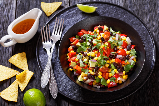 cowboy caviar, texas caviar with black bean, tomatoes, avocado, red bell pepper, corn, coriander  in black bowl on dark wooden table with nachos and dressing