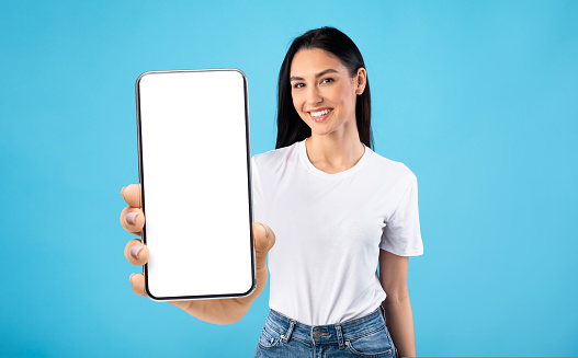 Happy young lady demonstrating big cellphone with white screen, showing space for your website or app design, standing over blue studio background, mockup