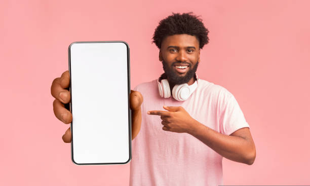 istock Mobile advertisement. Happy black man pointing at smartphone with empty white screen on pink background, mockup 1433053623
