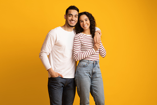 Portrait of happy embracing middle eastern couple posing over yellow studio background, cheerful young arab man and woman looking and smiling at camera, enjoying time together, copy space