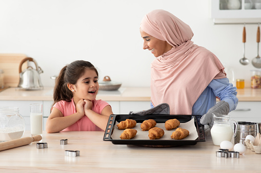 Excited Little Girl Looking At Muslim Mom With Tray Of Freshly Baked Croissants In Kitchen, Happy Islamic Family Mother And Daughter Enjoying Baking Together At Home, Ready To Taste Pastry, Closeup