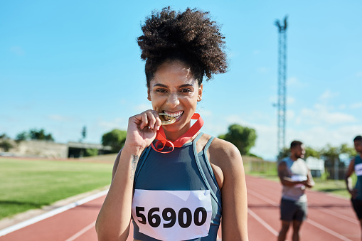 Female athlete, runner and black woman bite medal after win for sprinting or Olympic event at a stadium and track. Happy female champion enjoying celebration for winning award at sports competition