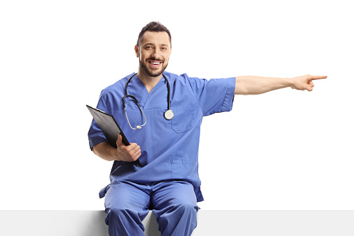 Male nurse in a blue uniform sitting on a blank panel and pointing to the side isolated on white background