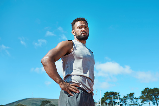 Fitness, black man and serious about exercise, health and wellness outside in blue sky nature for run, hike or cardio workout.  Portrait of athletic guy take break from exercising or training outdoor