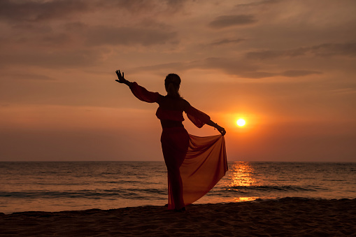 Silhouette middle aged woman dancing on tropical sea sunset. Female posing relaxing on summer beach at tropical background. Lady in beachwear long dress. Travel vacation concept. Copy text space