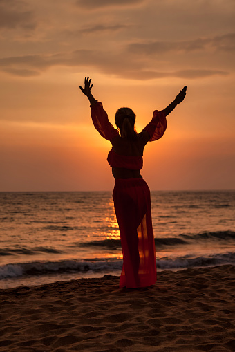 Silhouette middle aged woman arms raised on tropical sea sunset. Female posing relaxing on summer beach at tropical background. Lady in beachwear long dress. Travel vacation concept. Copy text space