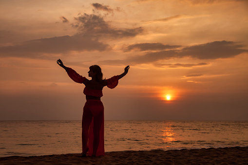 Silhouette middle aged woman arms raised posing on tropical sea sunset. Female relaxing on summer beach at tropical background. Lady in beachwear long dress. Travel vacation concept. Copy text space