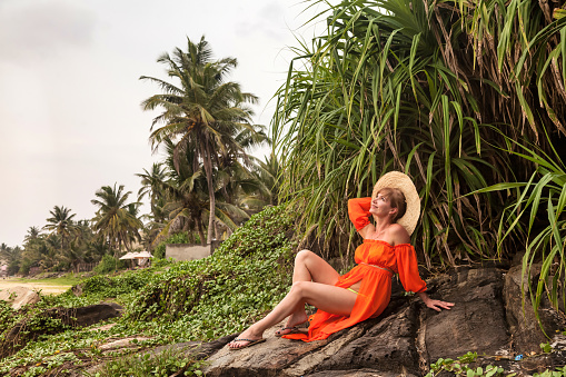 Middle aged woman in orange beachwear at tropical jungles background, looking at camera. Female with hat in tropical view with green palm trees in Sri Lanka. Travel vacation concept. Copy text space