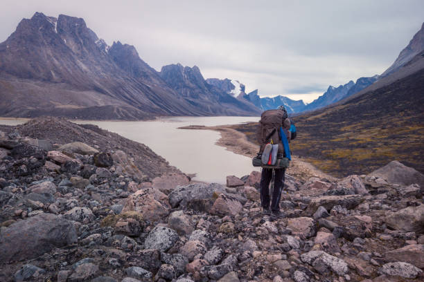 Hiker with a heavy backpack walking by a Summit lake in remote arctic valley on a cloudy day. Dramatic arctic landscape of Akshayuk Pass, Baffin Island, Canada. Mt. Thor in the distance. stock photo