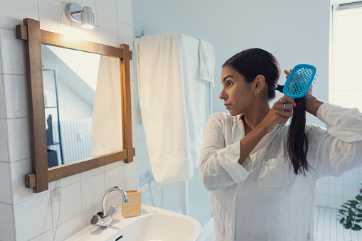 Woman in white pajamas brushing hair and looking in mirror at home