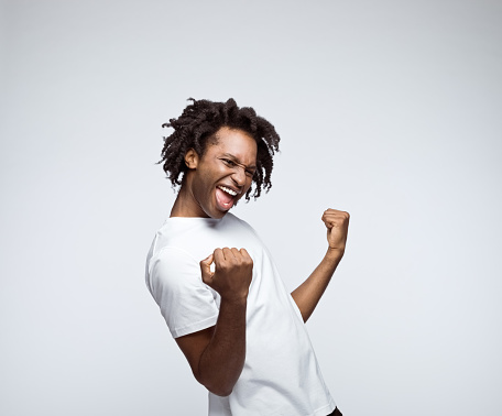 Happy afro american young man wearing white t-shirt, flexing first in winner gesture. Studio shot on grey background.
