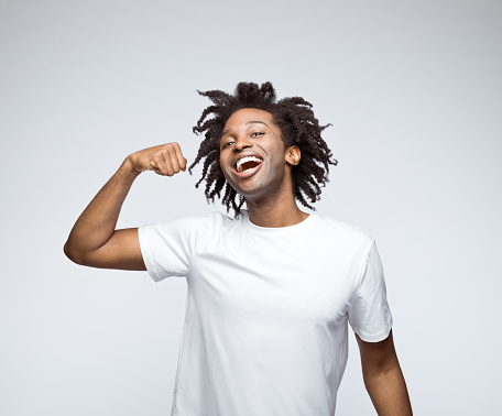 Afro american young man wearing white t-shirt, laughing at camera and flexing his muscles. Studio shot on grey background.