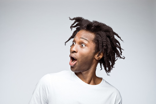 Surprised afro american young man wearing white t-shirt, looking away with mouth open. Studio shot on grey background.