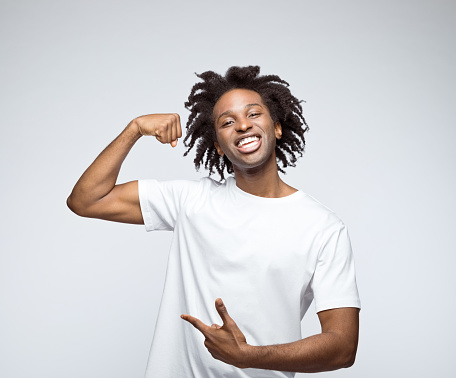 Excited afro american young man wearing white t-shirt, laughing at camera and flexing his muscles. Studio shot on grey background.