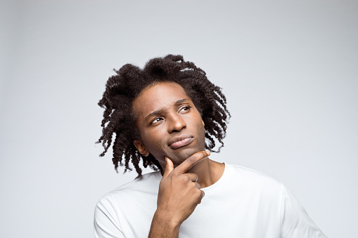 Headshot of afro american young man wearing white t-shirt, looking away with hand on chin. Studio shot on grey background.
