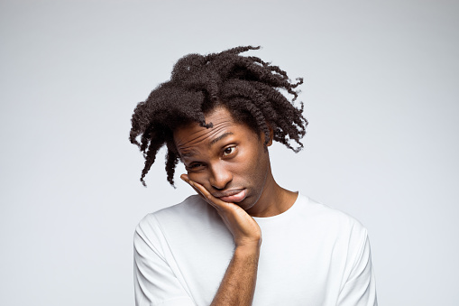 Displeased afro american young man wearing white t-shirt, looking away with hand on chin. Studio shot on grey background.