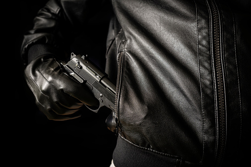 The concept of the crime of banditry. A dangerous shooter and a black pistol on a dark background. The hired killer is preparing to shoot. Pulls a firearm out of his jacket pocket