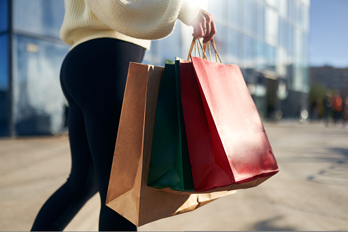 Close up of woman walking with shopping bags in hands in city from a mall. Low angle view of girl going with purchases from black friday sale. Female with full paper bags. Back view.