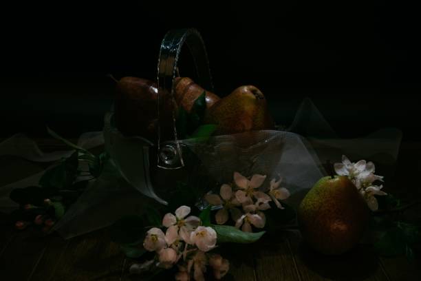 A glass basket with pears and white flowers on a table with a dark setting - still life photography