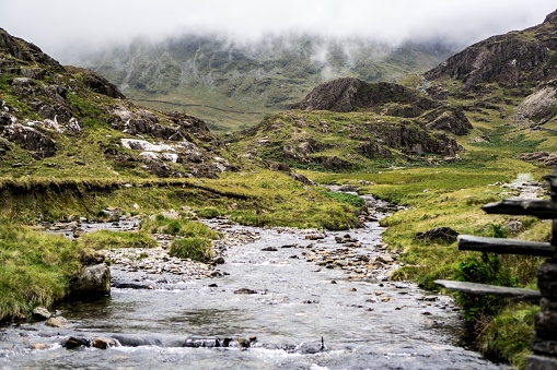 A scenic shot of a trickling river surrounded with green hills on a foggy overcast day