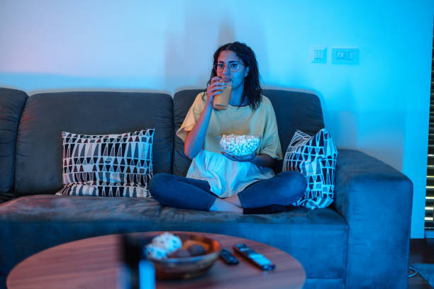 Young Latin Female Drinks Orange Juice While Watching TV. She Holds Popcorn stock photo