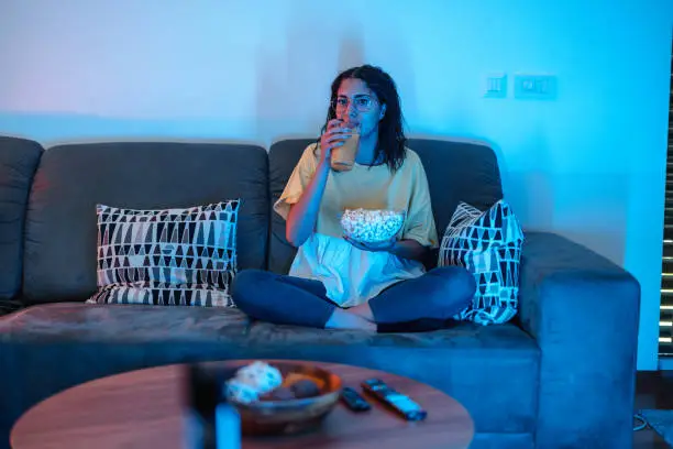 Young latin female with glasses is sitting on her sofa, drinking natural orange juice and holding popcorn while watching her favourite show.