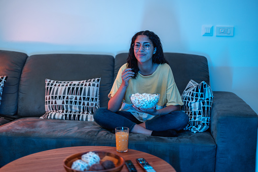 Young Hispanic female is sitting on a comfortable grey sofa, watching a movie. She is eating popcorn.