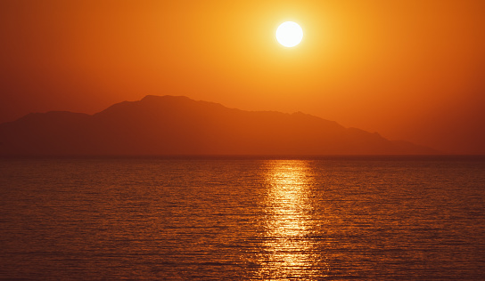beautiful orange tone sunset sunrise over the sea or ocean, big sun above mountain hill, sun specular reflection in water, scenic landscape, travelling around the world