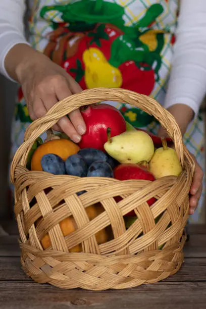 Wicker basket of fresh autumn fruit and a young woman with an apron standing in the background. Vertical shot, a close-up.
