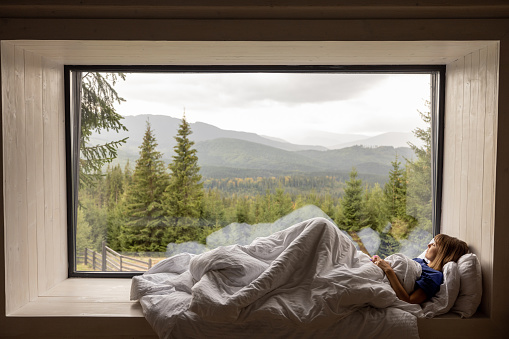 Woman lying on window sill covered with blanket, enjoying great view on mountains and pine forest during vacation in house on nature