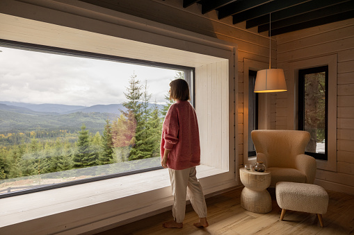 Woman enjoys scenic view on mountains while resting in wooden house on nature. Recreation and home coziness concept